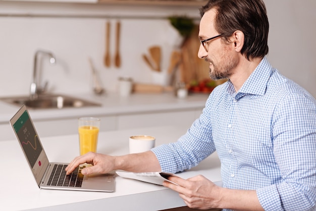 Full of inspiration. Smiling satisfied sincere self employed businessman sitting at home and while using laptop and reading new