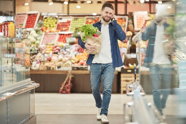 Full length of adult bearded man calling by smartphone while grocery shopping in supermarket