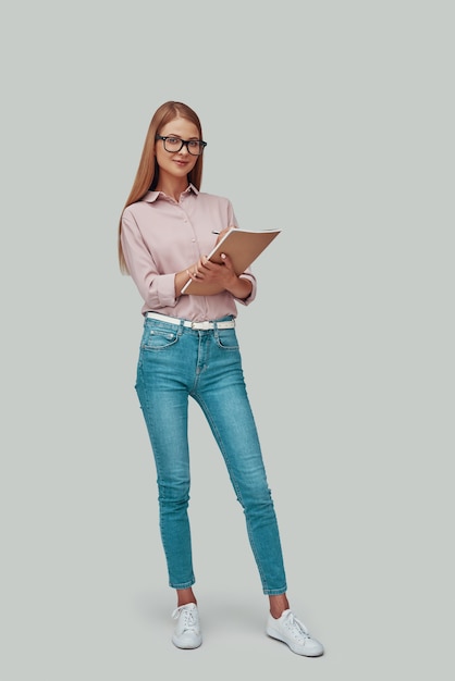 Full length of attractive young woman writing something down and smiling while standing against grey background