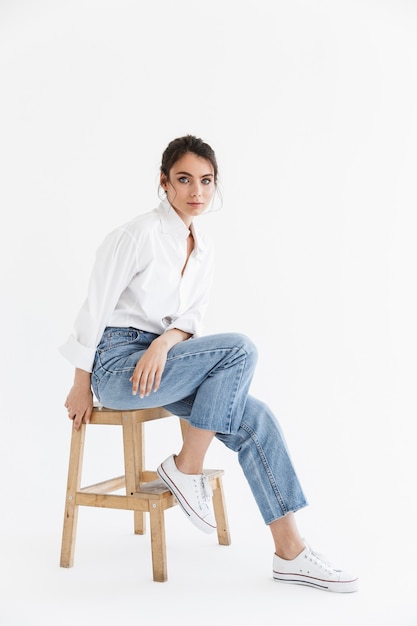 Full length of a beautiful sensual young woman with long curly brunette hair wearing white shirt sitting on a chair isolated over white wall, posing