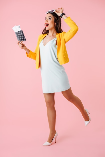 Full length of a beautiful young woman wearing colorful clothes standing isolated over pink, showing passport with flight tickets