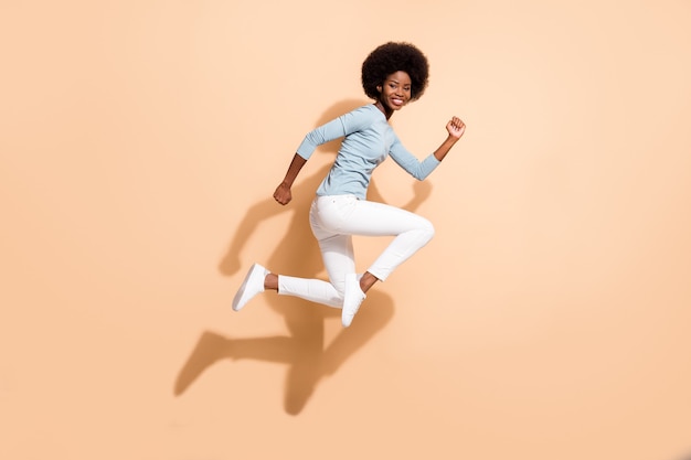 Full length body size side profile photo of pretty dark skinned active girl jumping high running fast smiling isolated on beige color background