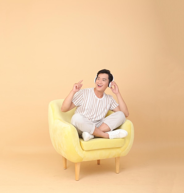 Full length body stock photo of a handsome Asian young man sit on yellow sofa chair isolated on beige background Concept for app user high tech gadgets or Internet promotion