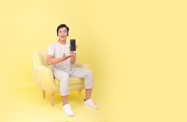 Full length body stock photo of a handsome Asian young man sit on yellow sofa chair isolated on yellow background show smart phone screen Concept for app user high tech gadgets or Internet promotion