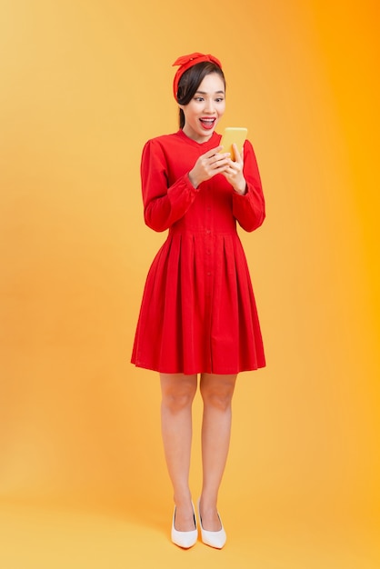 Full length body of surprised young Asian woman reading news while standing over orange background.