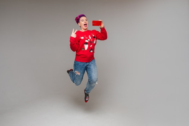 Full length of carefree jovial woman with dyed purple hair in red sweater jumping while making selfie on her smartphone showing peace gesture with two fingers