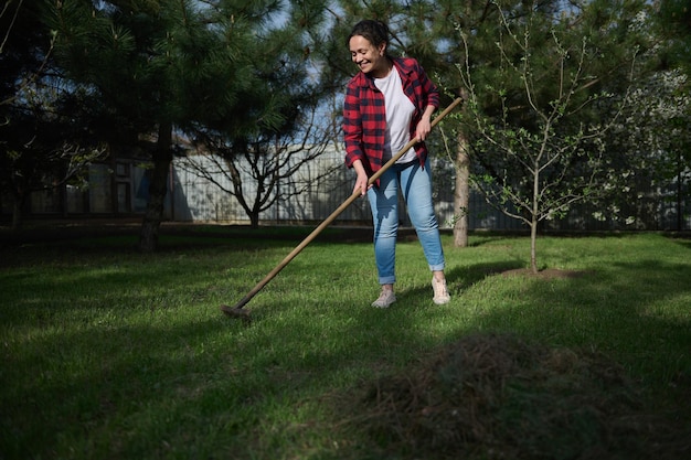 Full length a charming Hispanic woman gardener cleaning the local area and raking grass against blossoming fruit tree background on a sunny spring day