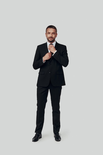 Full length of charming young man in full suit adjusting tie while standing against grey background