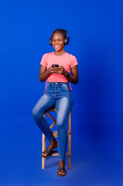 Full length of cheerful young black woman with headphones and cellphone listening to music or audio book sitting on chair