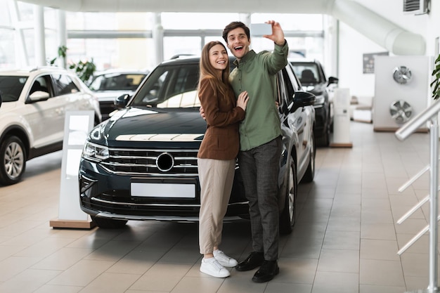 Full length of cheery young couple taking selfie on mobile phone near their new car at auto dealership copy space