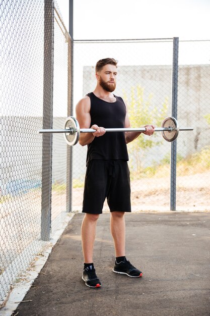 Full length of concentrated young man athlete working out with barbell outdoors