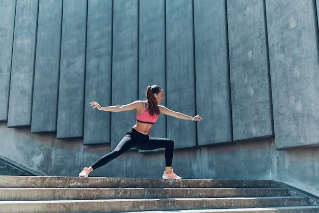 Full length of confident young woman in sports clothing doing stretching exercises outdoors