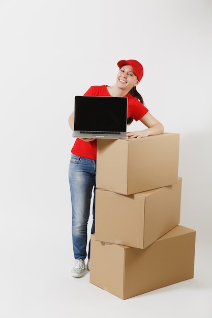 Full length of delivery woman in red cap, t-shirt isolated on white background. Female courier near empty cardboard boxes, laptop pc computer, blank black empty screen. Receiving package. Copy space.