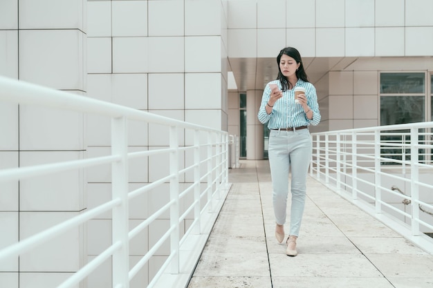 full length of elegant young office lady walking in sky corridor outdoor in skyscraper. confident female business people using cellphone and holding takeaway coffee enjoy sunshine on bridge.