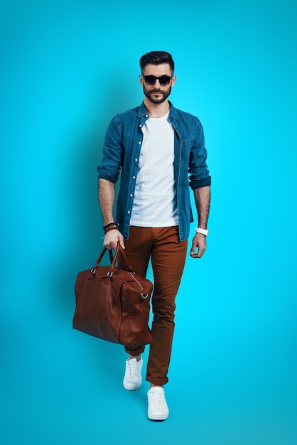 Full length of fashionable young man smiling and looking at camera while walking against blue wall