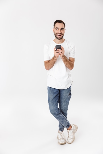 Full length of a handsome cheerful man wearing blank t-shirt standing isolated over white wall, using mobile phone