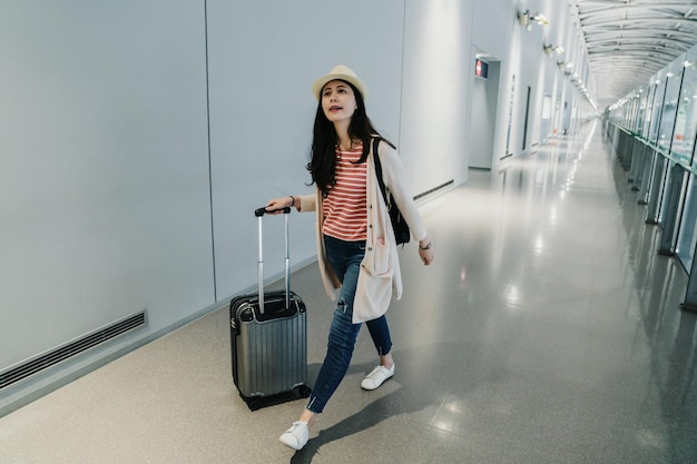 full length happy japanese girl traveler keeping luggages while walking in airport going to boarding gate lounge. self guided trip lifestyle concept. chinese female backpacker in hat with suitcase.