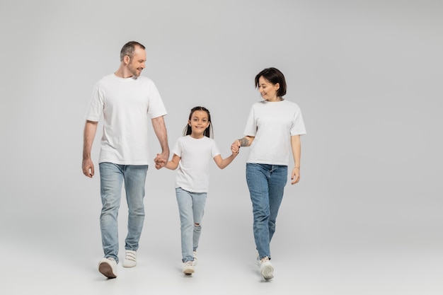 full length of happy parents and kid in white tshirts and blue denim jeans holding hands and