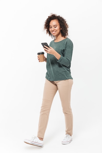 Full length of a happy young african woman wearing casual clothes isolated over white wall, using mobile phone, drinking takeaway coffee