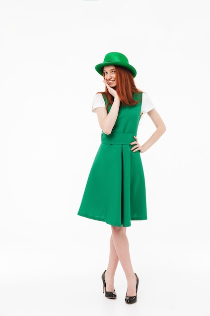 Full length of a happy young redheaded girl wearing green hat, celebrating St Patricks's Day isolated over white wall, posing