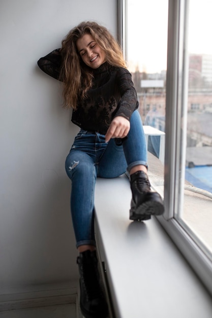 Full length image of a cheerful gorgeous young woman with curly hair wear black blouse and jeans posing near windows