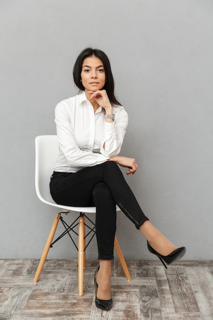 Full length image of elegant businesswoman 30s in formal wear sitting on chair in office, isolated over gray background