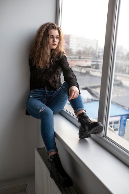 Full length image of a gorgeous young woman with curly hair wear black blouse and jeans posing near windows