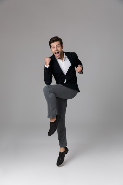 Full length image of handsome businessman 30s in formal suit yelling and clenching fists, isolated over gray wall