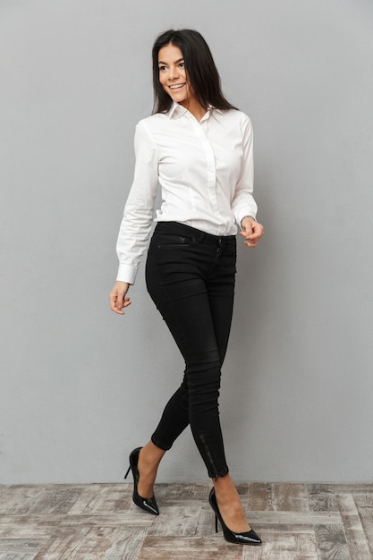 Full length image of happy businesswoman in white shirt and black trousers walking and looking aside with smile, isolated over gray background