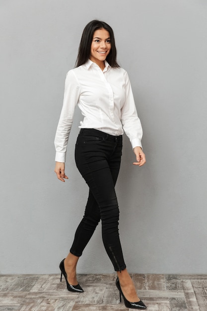 Full length image of smiling businesslike woman in white shirt and black trousers walking and looking aside, isolated over gray background