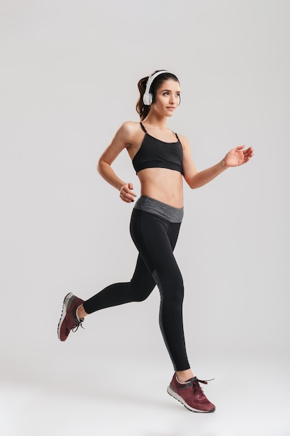 Full-length image of sporty woman in tracksuit running and listening to music with wireless headphones, isolated over gray wall