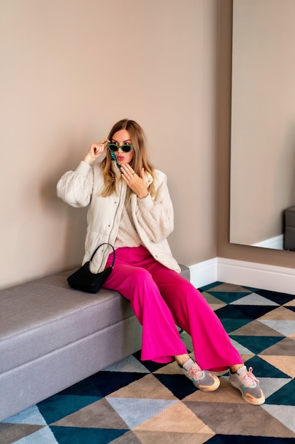 Full length image of trendy blonde woman posing at luxury hotel, wearing warm colorful casual outfit