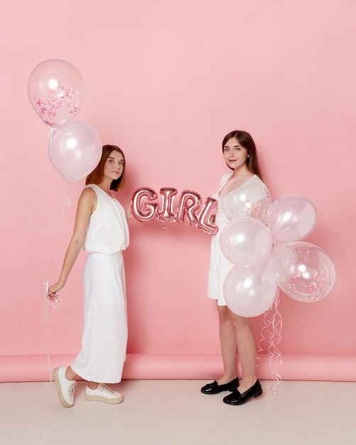 Full length image of a two of young friends dressed in a white holdings a balloons over pink background