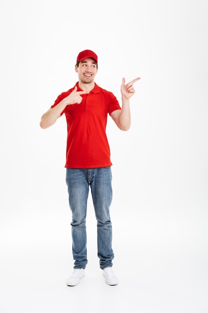 Full length image of young courier in red uniform presenting copyspace text or product with pointing fingers aside and smiling broadly, isolated over white space