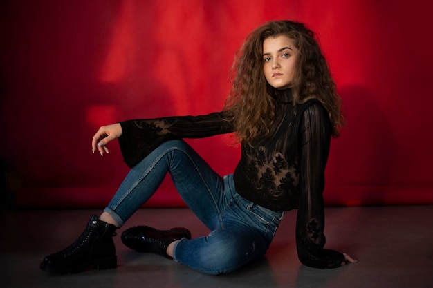 Full length image of a young woman with makeup and curly hair dressed black blouse and jeans isolated red background