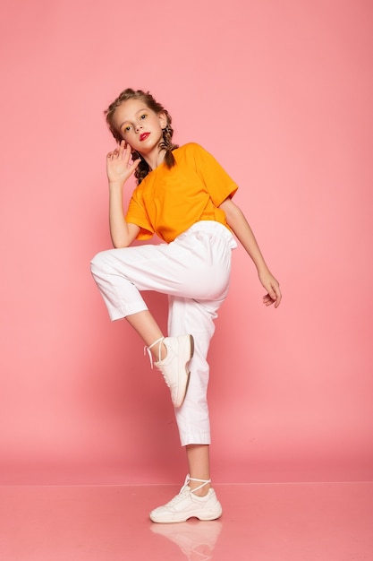 Full length little girl on pink studio wall. wearing an orange T-shirt and white pants and white sneakers.  