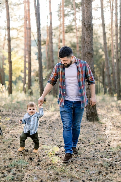 Full length outdoor portrait of handsome bearded young man father, walking in pine autumn forest together with his little adorable child son