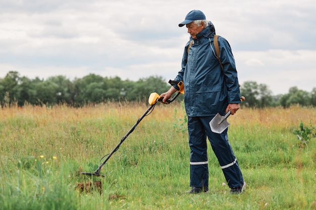 Full length outdoor portrait of numismatist in jacket and cap, holding in hands shovel and metal detector, looking for gold or historical artifacts in meadow.