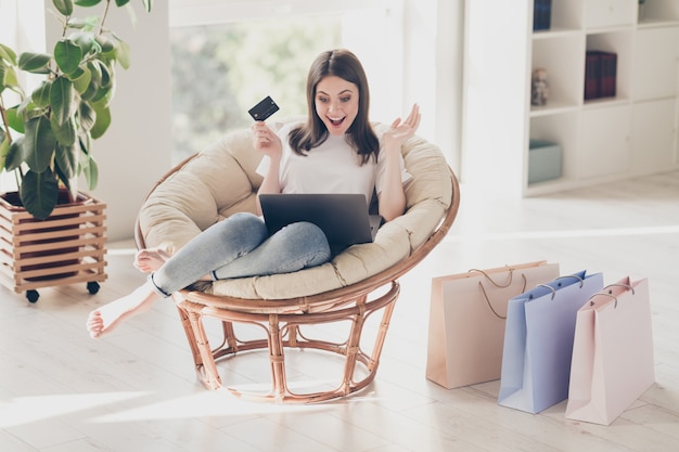 Photo full length photo of amazed girl sit chair look laptop monitor hold credit card in house indoors shopping sales concept