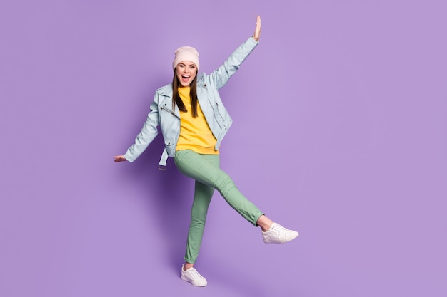 Full length photo of attractive funny lady spring weather nice day street clothes good mood walk raise leg arms wear casual hat modern jacket pants shoes isolated purple color background