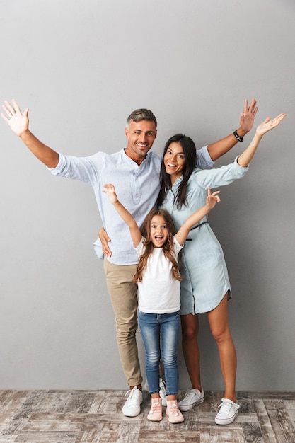 Full length photo of beautiful caucasian family woman and man with little girl smiling and hugging together, isolated over gray