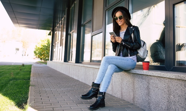 Full-length photo of fancy woman in a black hat and sunglasses, who is sitting near a modern building, checking feed in her smartphone and drinking coffee