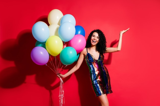 Full length photo of gorgeous girl hold many balloons raise arm palm wear glossy short dress isolated bright red color background