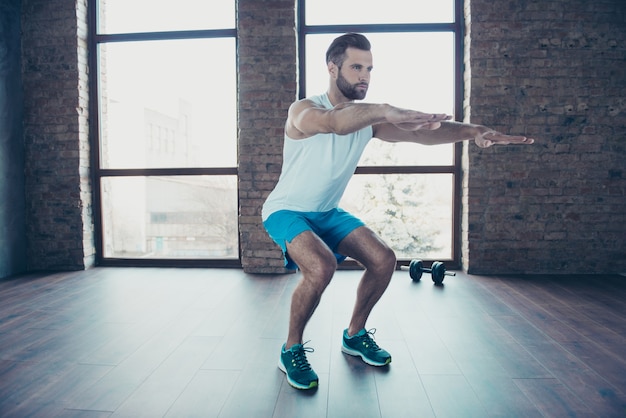 Full length photo of handsome trainer guy doing static squats concentrated sportswear tank-top shorts sneakers training home  near windows indoors