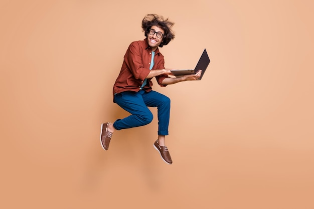 Full length photo portrait of excited guy jumping up with laptop in hands isolated on pastel beige colored background