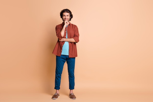 Full length photo of smart guy look camera hand chin wear brown t-shirt blue pants footwear isolated beige color background