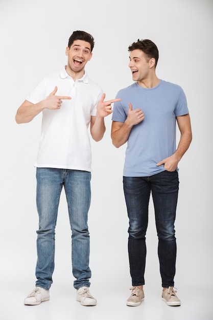 Full length photo of two handsome men 30s wearing casual t-shirt and jeans smiling and gesturing fingers on each other, isolated over white background