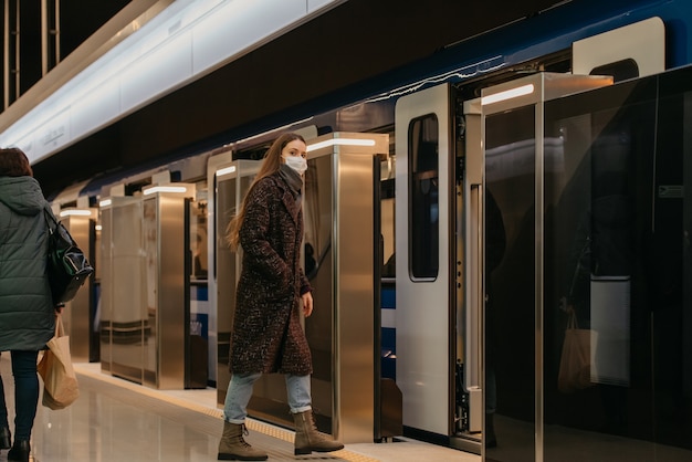 A full-length photo of a woman in a medical face mask to avoid the spread of coronavirus who is entering the modern subway car. A girl in a surgical mask is keeping social distance on a metro station.