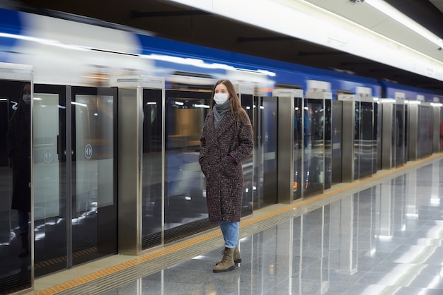 A full-length photo of a woman in a medical face mask waiting for an arriving modern train on the subway