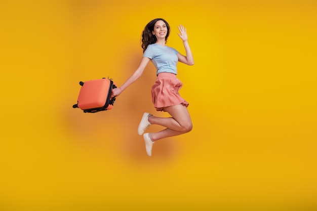 Full length photo of young attractive woman happy positive smile suitcase trip wave hello isolated over yellow color background